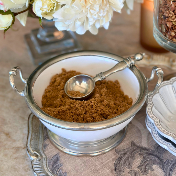 White bowl with brown powder and a silver scoop on a decorative tablecloth.