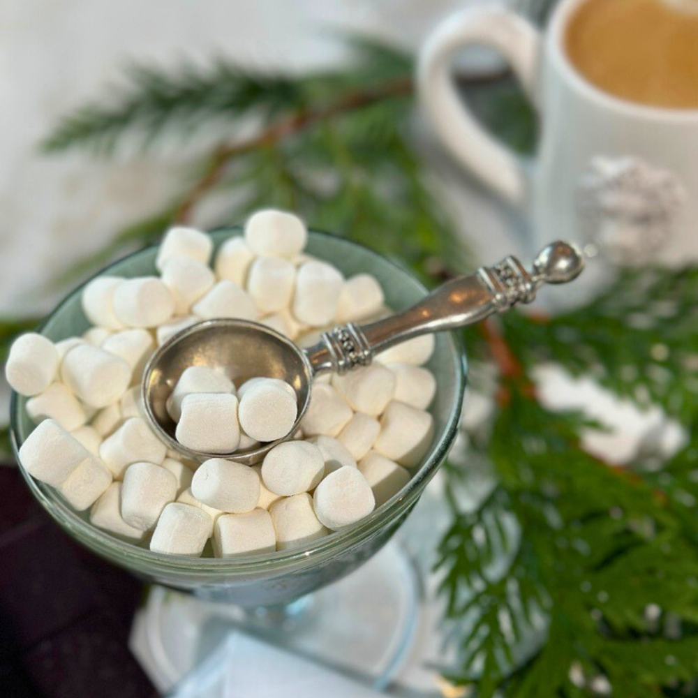 Glass dish with marshmallows and a silver scoop, with a blurred coffee cup in the background.