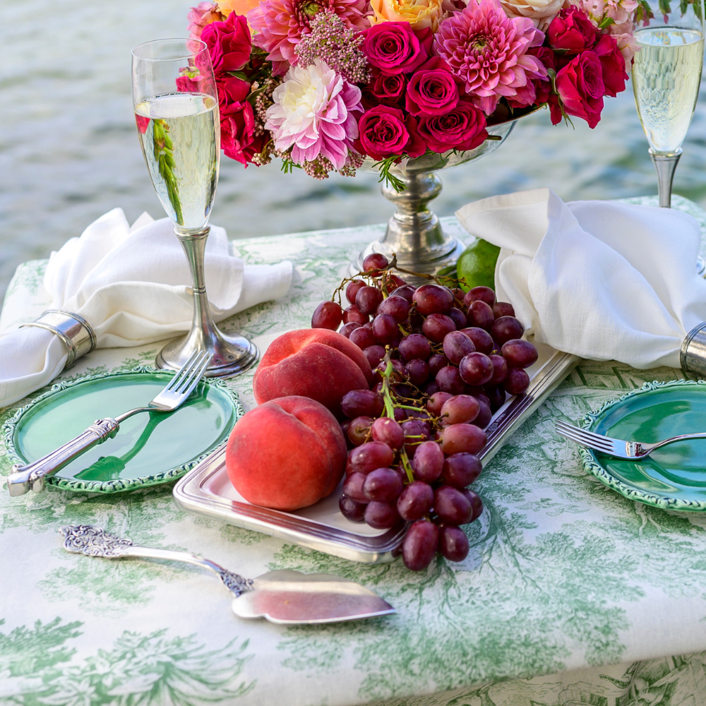 Table setting with fruits, flowers, and champagne glasses by a body of water.