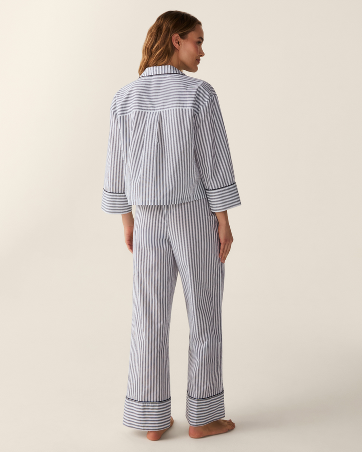 A woman stands barefoot from the back wearing a matching striped pajama set with a long-sleeve button-front top and wide-leg pants, in a light gray and white vertical stripe pattern against a warm off-white studio background.