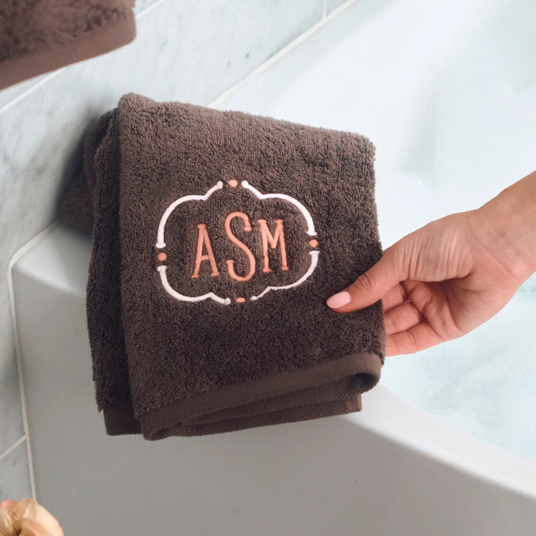 brown hand towel sitting on edge of bath towel with embroidery