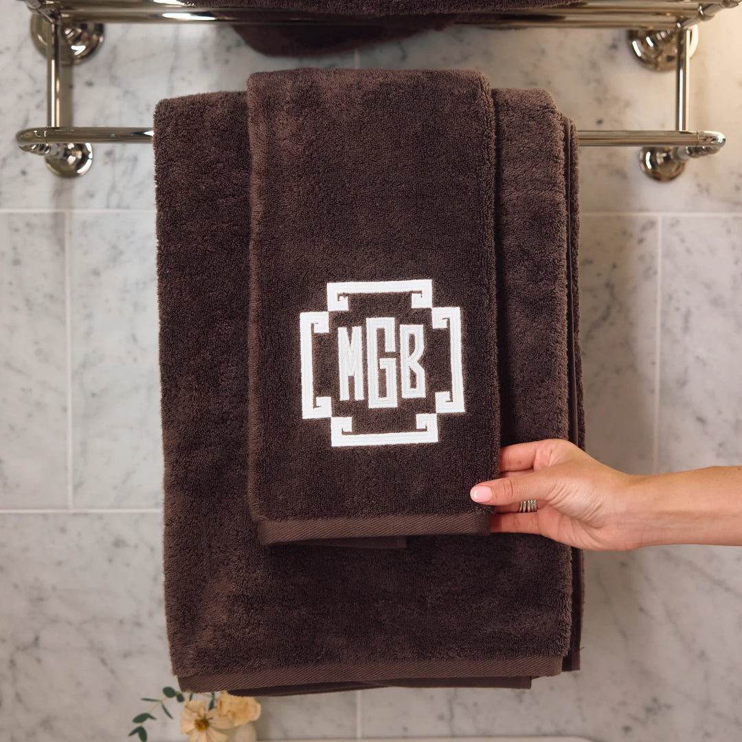 A dark brown plush towel set hanging on a chrome towel bar against a light marble tile wall. The top towel features a white geometric monogram design containing the letters 'MGB'.