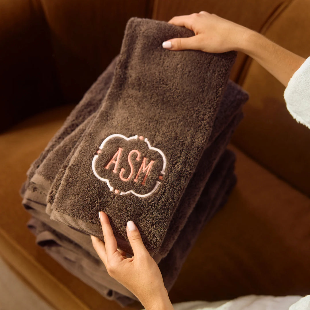 A stack of dark brown plush towels held by hands, with the top towel featuring an embroidered monogram design that reads 'ASM' inside a decorative border.