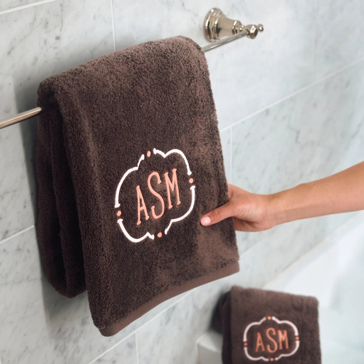 A dark brown plush bath towel with a white and peach embroidered monogram 'ASM' hangs on a chrome towel bar in a tiled bathroom. A hand is holding the towel, and a smaller matching towel is visible at the bottom right.