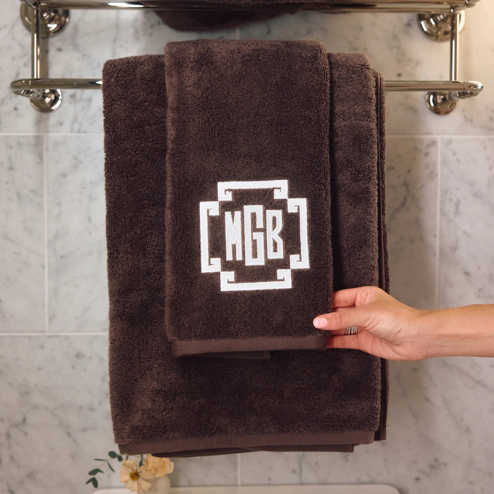 A dark brown plush towel set hanging on a chrome towel rack against a light marble tile wall. The set includes a larger bath towel and a smaller hand towel, both with a white geometric monogram design reading 'MGB'.