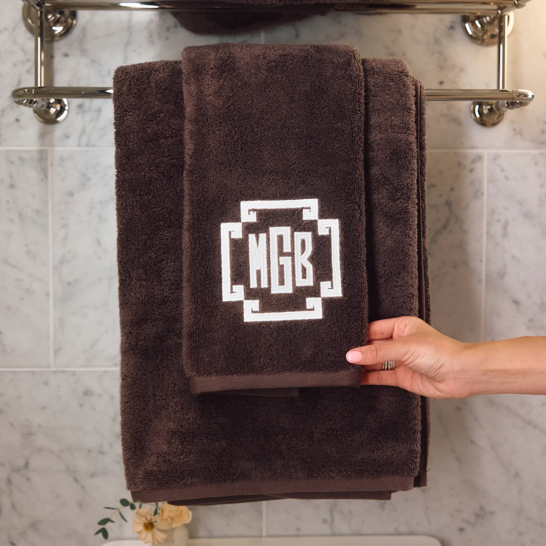 A dark brown plush towel set hanging on a chrome towel rack against a light marble tile wall. The set includes a larger bath towel and a smaller hand towel, both with a white geometric monogram design reading 'MGB'.