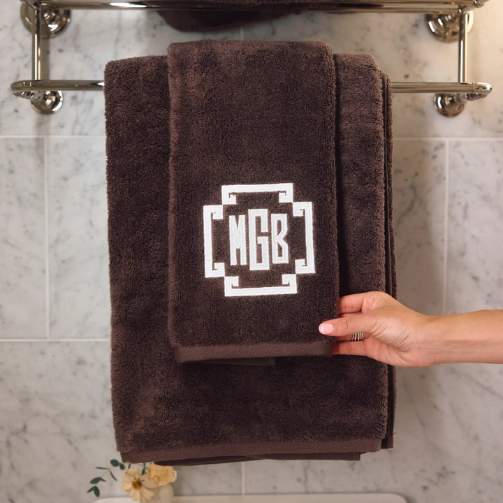 A dark brown plush towel set hanging on a chrome towel rack against a light marble tile wall. The set includes a larger bath towel and a smaller hand towel, both with a white geometric monogram design reading 'MGB'.