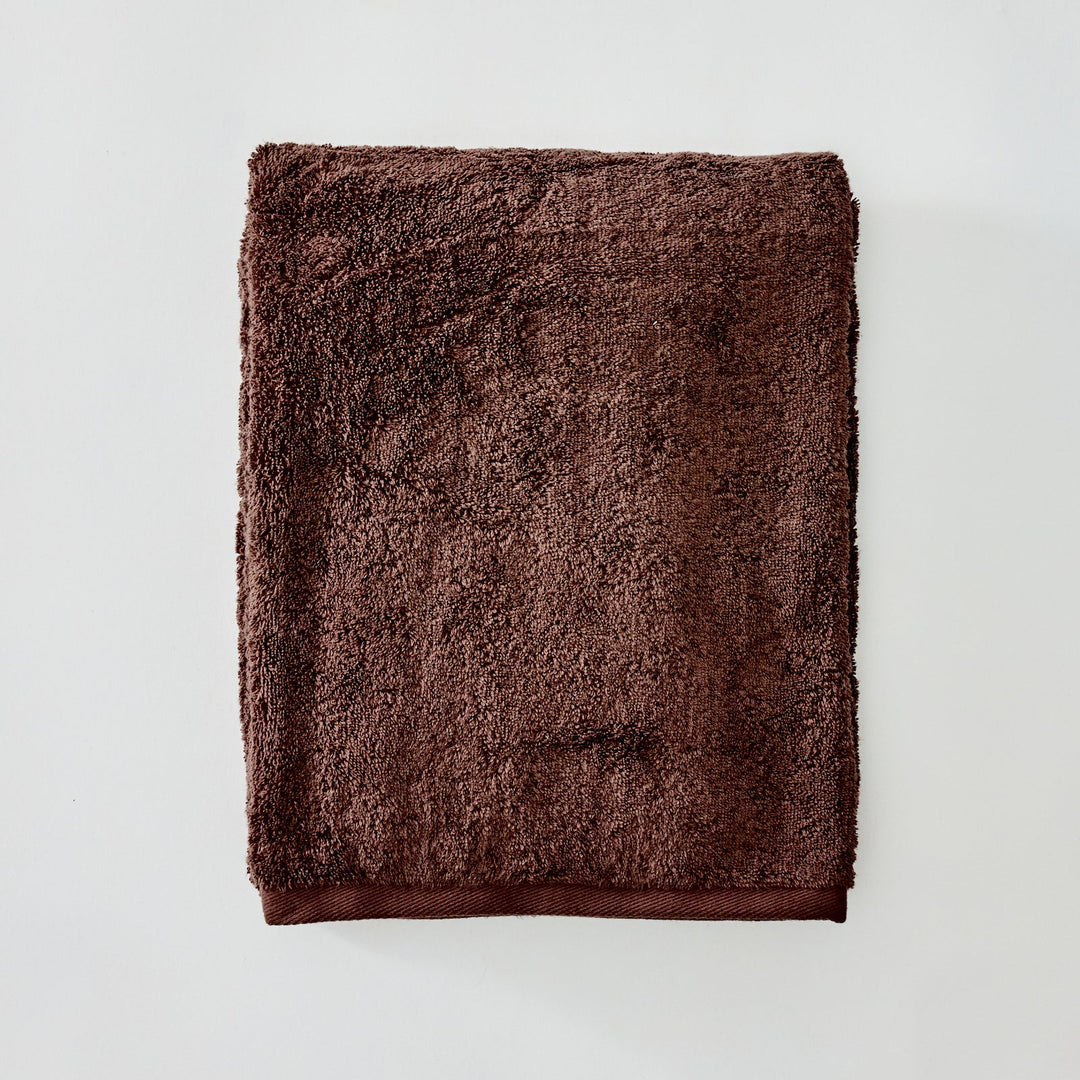 A folded brown terry cloth towel with a thick, plush texture and a matching solid brown border, displayed flat against a light gray background.