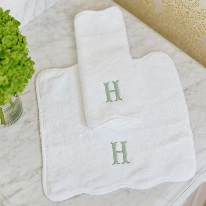 A set of white baby bath towels, including a large square bath mat and a smaller folded towel, both embroidered with a green letter 'H', displayed on a light marble surface next to a green hydrangea.