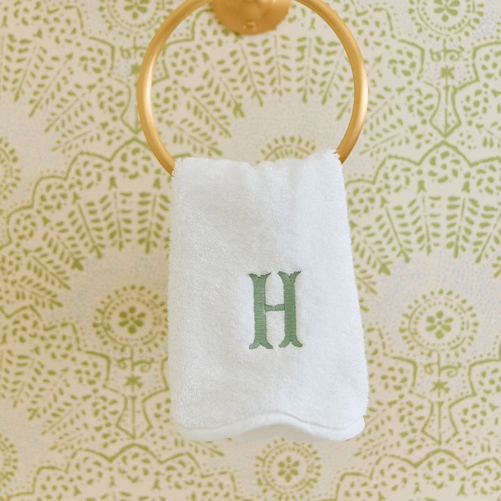 A white hand towel with a green embroidered letter 'H' hangs from a gold ring on a patterned cream and green wall.