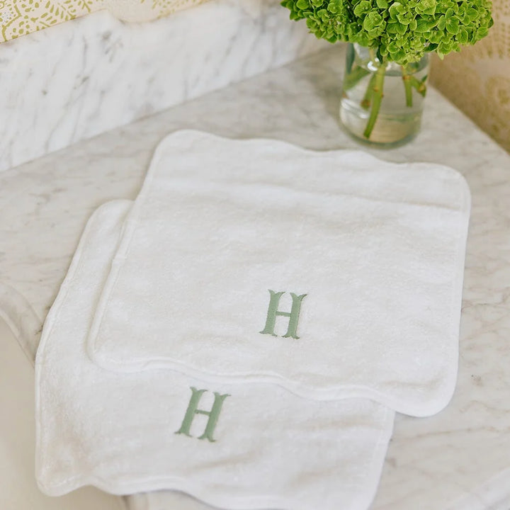 Two white cotton baby washcloths with scalloped edges and embroidered green letter H monogram, displayed on a marble surface next to a glass vase of green hydrangeas.