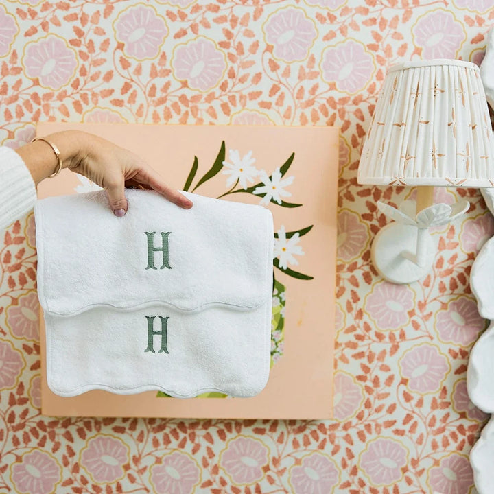 A hand holds a white folded towel or napkin with a green embroidered letter H, placed on top of a peach floral box against a pink and white patterned wallpaper background.