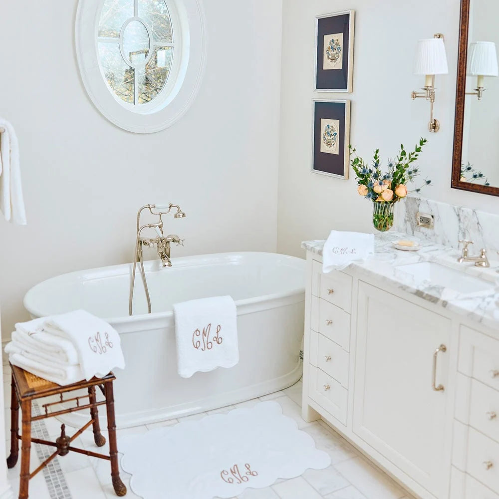 A bright, elegant bathroom featuring a white freestanding bathtub with antique brass fixtures, a matching white marble vanity, and personalized white towels and bath mats with gold monogram lettering.
