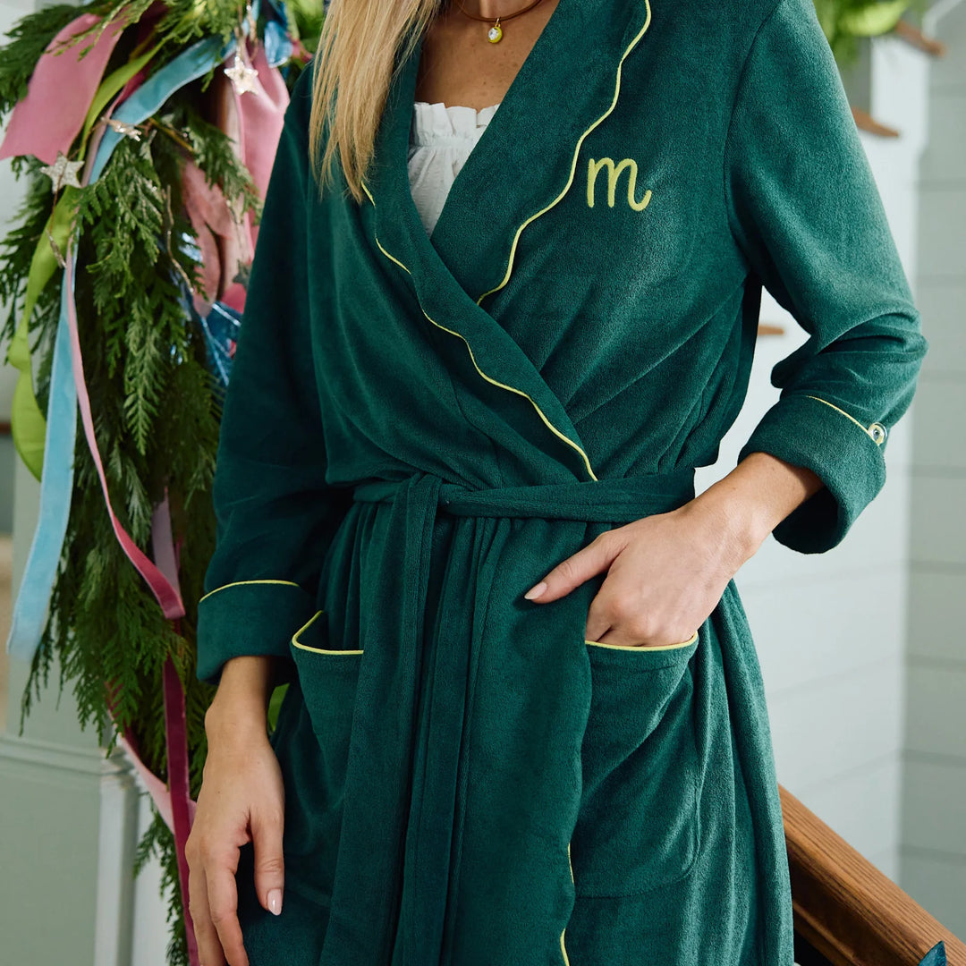 A woman wearing a dark green plush bathrobe with yellow piping and a yellow embroidered monogram 'm' on the chest. The robe has a tie belt and front pockets, styled over a white top in a home setting with holiday greenery in the background.