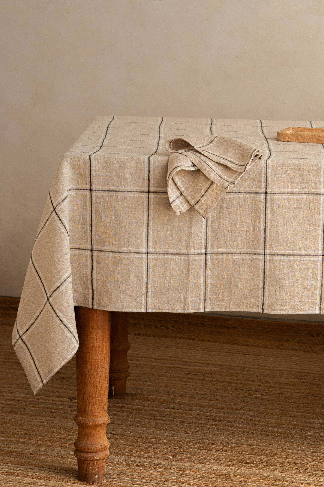 A beige plaid tablecloth draped over a wooden table, with a matching folded napkin on top, set against a neutral beige wall and woven jute floor.