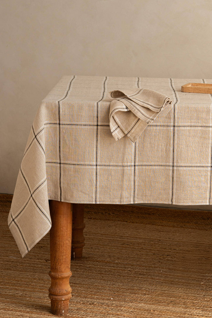 A beige plaid tablecloth draped over a wooden table, with a matching folded napkin on top, set against a neutral beige wall and woven jute floor.
