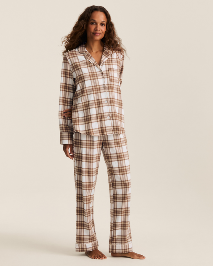 A woman modeling a two-piece pajama set with a brown and beige plaid pattern, standing against a solid light beige background.