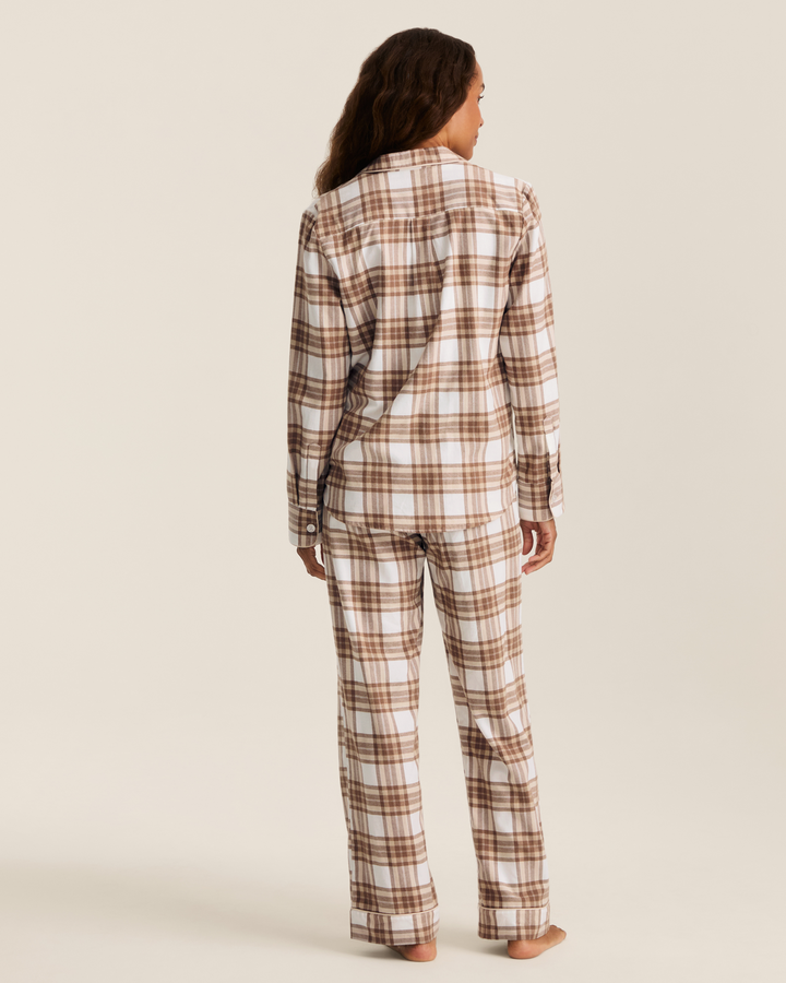 A woman shown from the back wearing a matching plaid pajama set with a long-sleeve button-up shirt and pants, in brown and cream tones, against a plain light beige studio background.