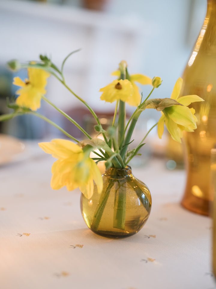 Yellow flowers arranged in a small round translucent amber glass vase on a white tablecloth, with a softly blurred indoor background and a larger amber bottle at the right edge.