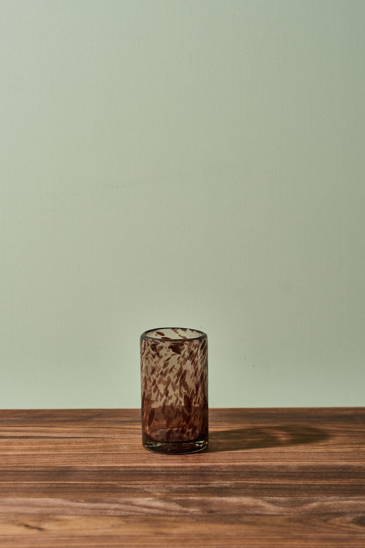 A small amber-brown textured glass tumbler with a swirled pattern, standing on a wooden tabletop against a pale sage green wall.
