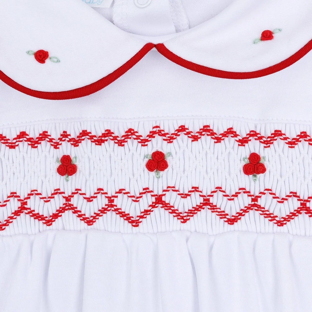 Close-up of a white baby smocked dress featuring red embroidered patterns, a Peter Pan collar, and small red flower details.