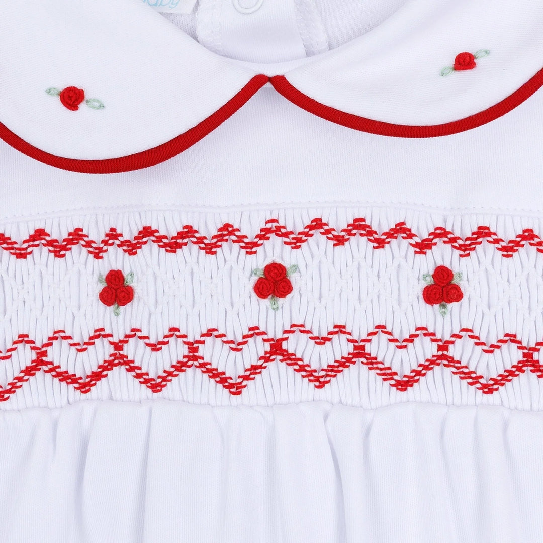 Close-up of a white baby smocked dress featuring red embroidered patterns, a Peter Pan collar, and small red flower details.