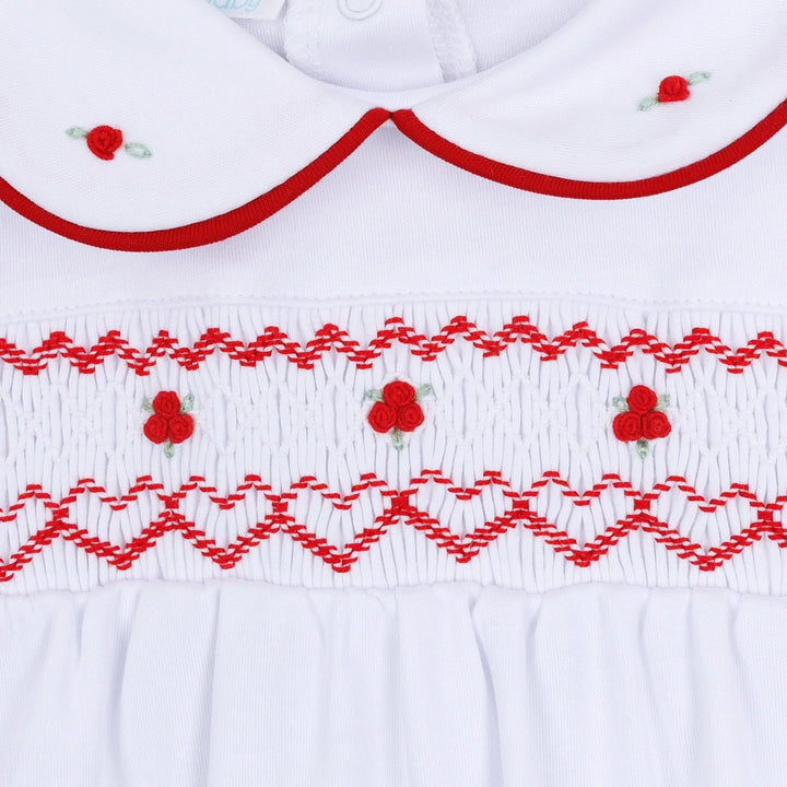 Close-up of a white baby smocked dress featuring red embroidered patterns, a Peter Pan collar, and small red flower details.