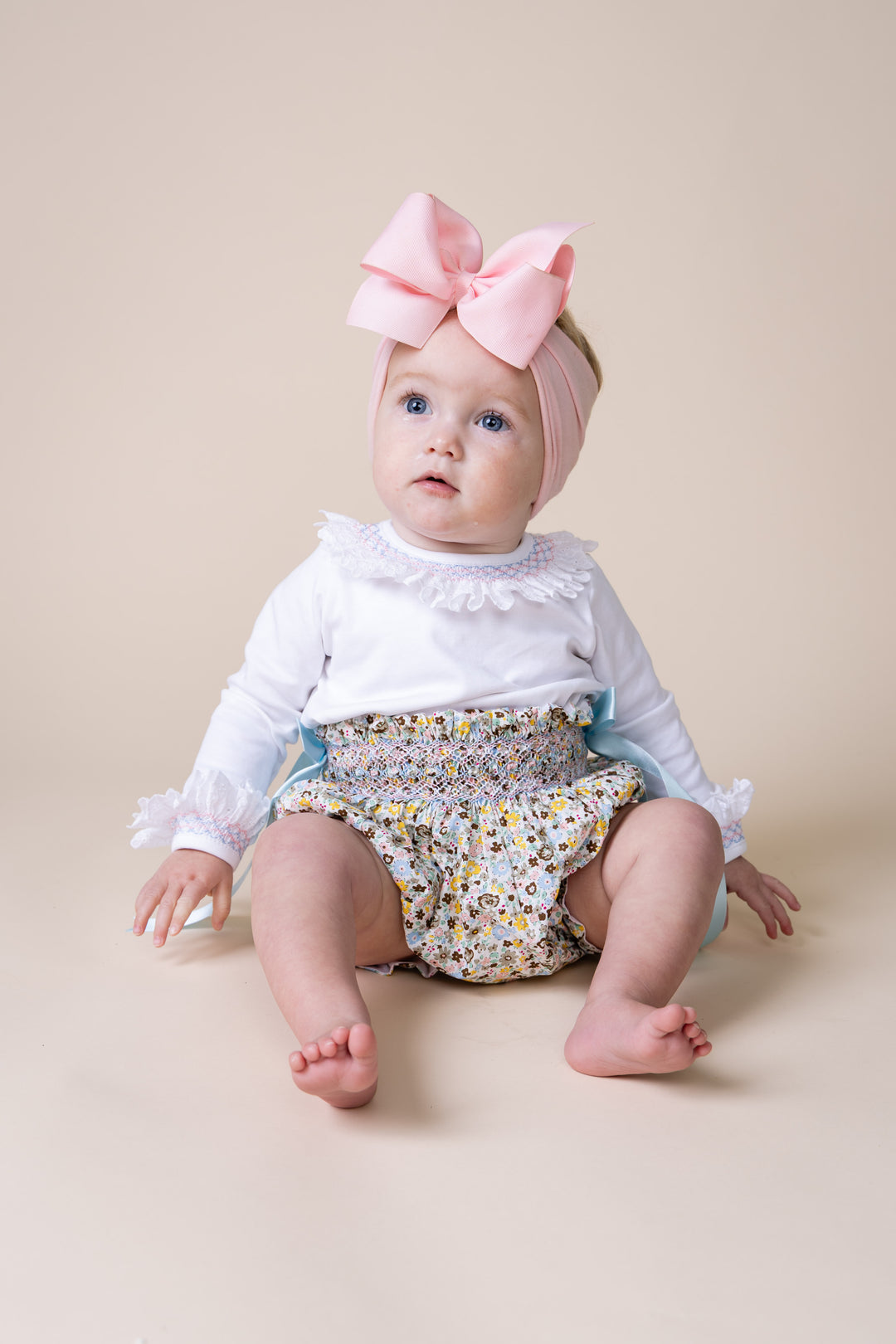 A baby girl sitting on a beige studio background, wearing a light pink headband with a large matching bow, a white long-sleeve top with ruffles, and a floral smocked diaper cover.