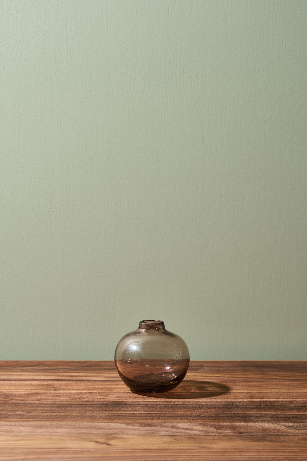 A small, round, smoky brown glass vase sits on a wooden tabletop against a solid sage green wall.
