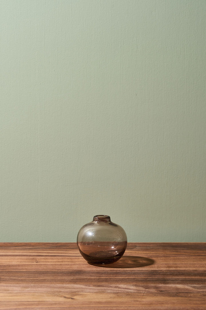 A small, round, smoky brown glass vase sits on a wooden tabletop against a solid sage green wall.