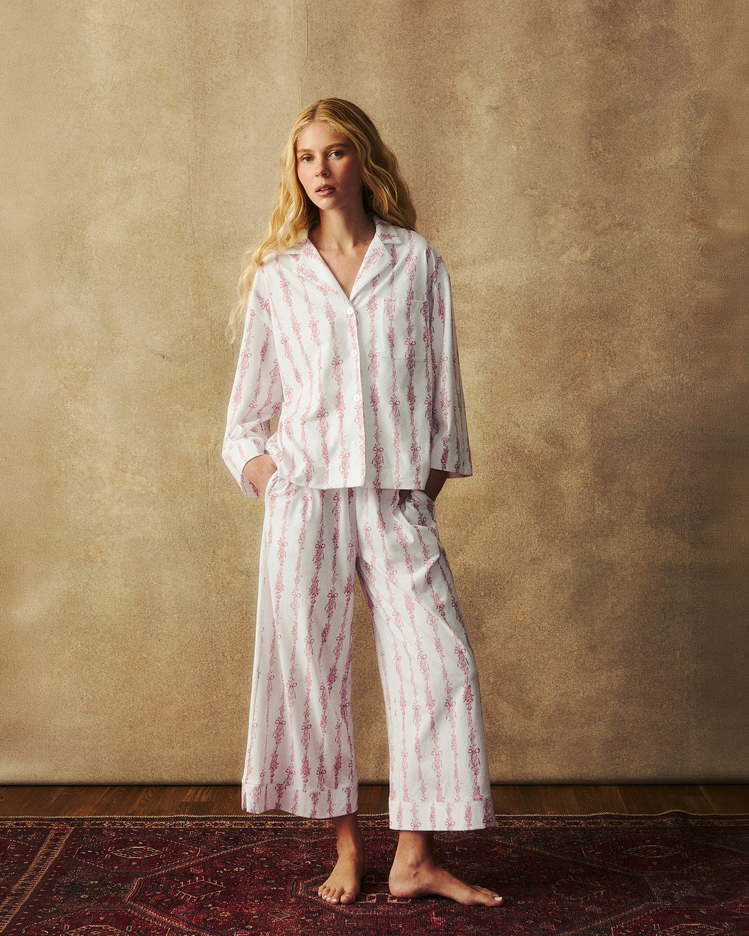 A woman stands barefoot indoors wearing a matching pajama set with a white base and vertical pink floral stripes. The set includes a long-sleeve button-front top and wide-leg pants, posed against a textured warm beige wall on a dark patterned rug.