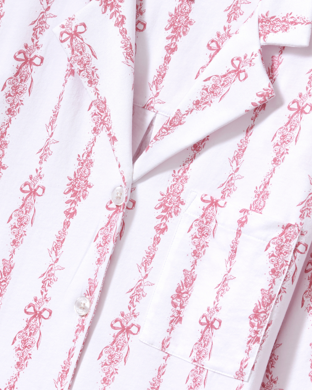 Close-up of a white button-front garment with a pink toile-style floral and bow print, showing part of the collar, front placket, and chest pocket.