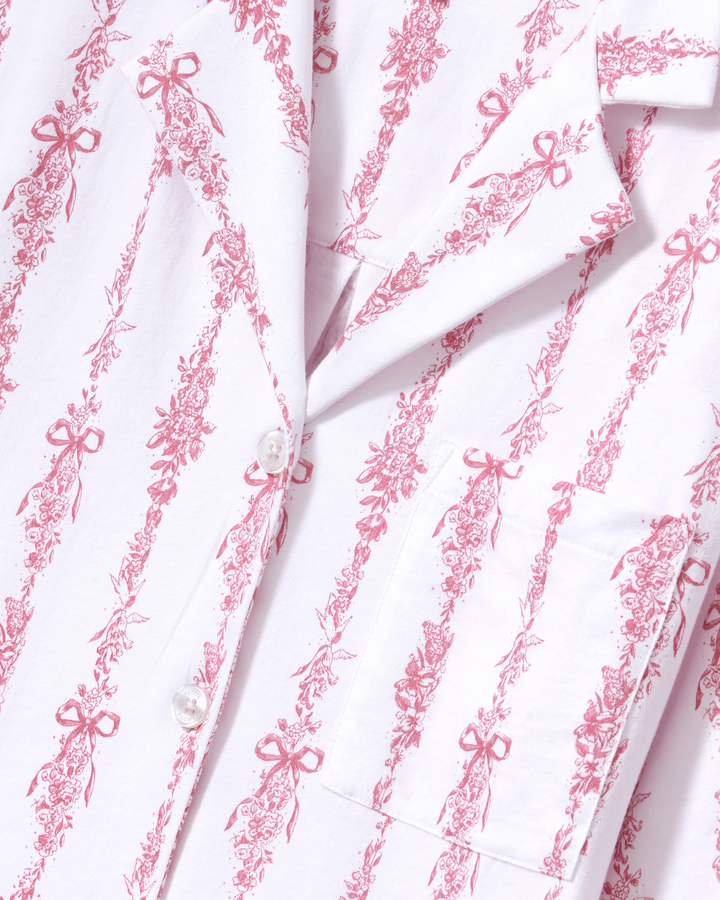 Close-up of a white button-front garment with a pink toile-style floral and bow print, showing part of the collar, front placket, and chest pocket.
