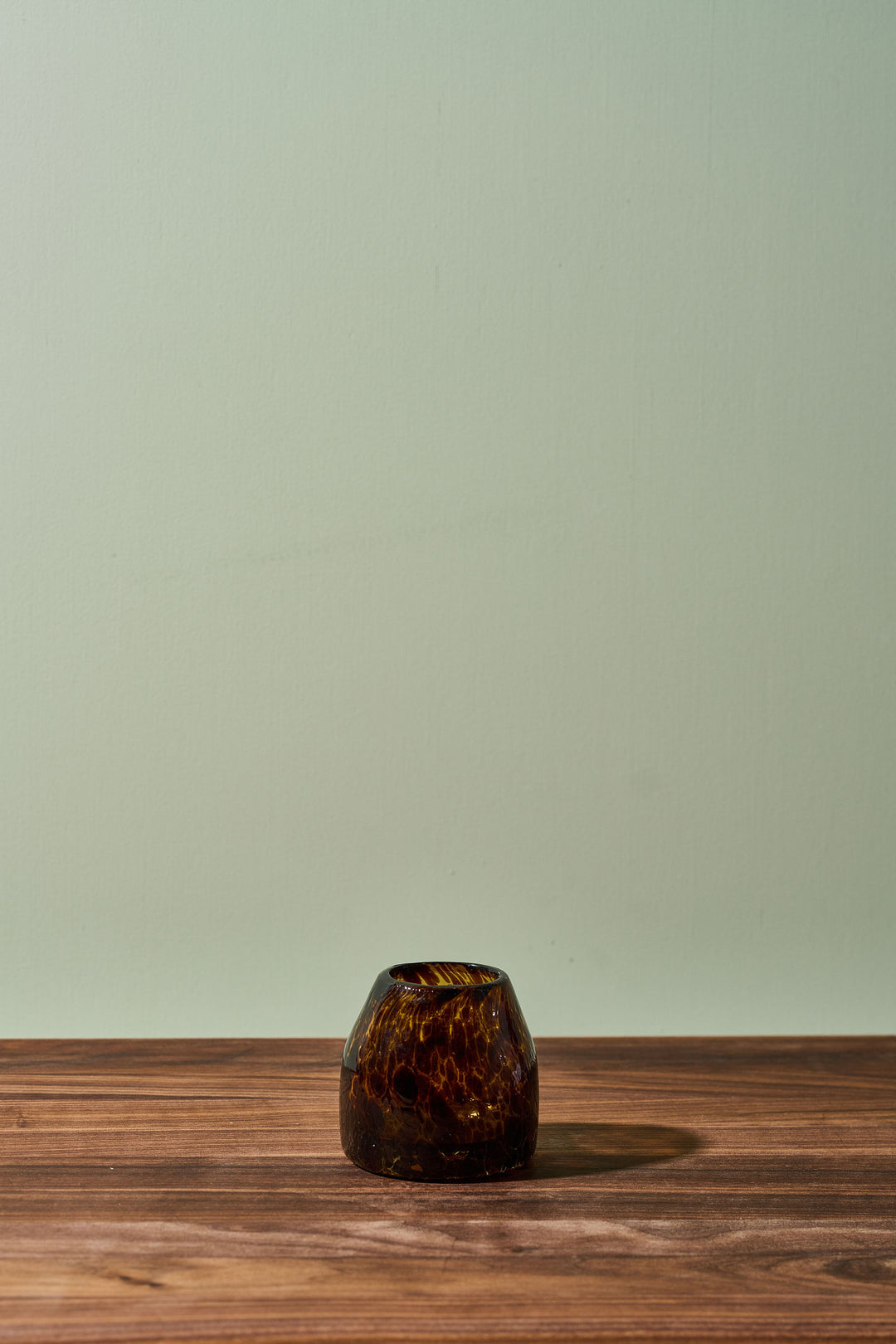 A small amber-brown marbled glass candle holder or vase sits on a wooden tabletop against a pale sage green wall.