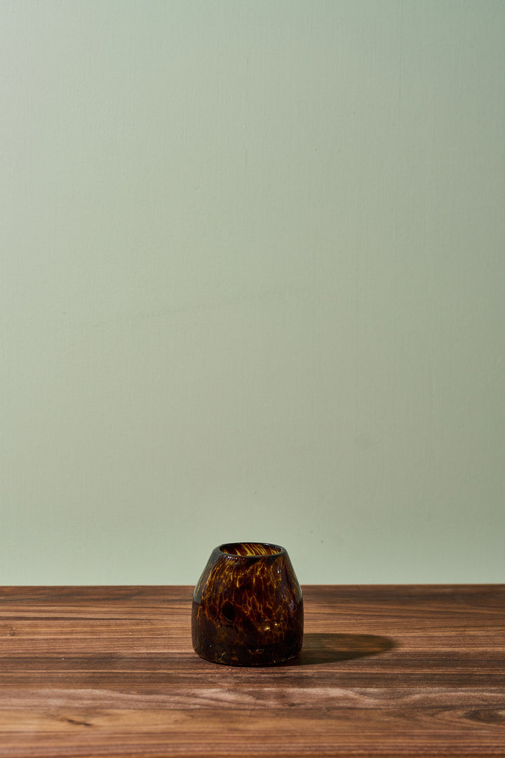 A small amber-brown marbled glass candle holder or vase sits on a wooden tabletop against a pale sage green wall.